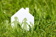 A paper-cutout of a family in front of a paper cutout of a house, in a rural field of grass.